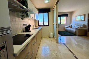 A modern kitchen with a built-in stove, a sink, and a window. There is a sliding door leading to a living area with a sofa and a small table. - Casa Ribera Beach by Rental Hero (Narejos, Los)
