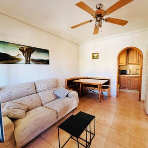 Un salon comprenant un canapé, une table à manger, un ventilateur de plafond et un coin cuisine avec des meubles en bois.