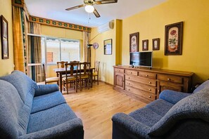 Ein Wohnzimmer mit zwei Sofas, einem Esstisch mit Stühlen, einem Fernseher auf einem Holzschrank und einem Deckenventilator.