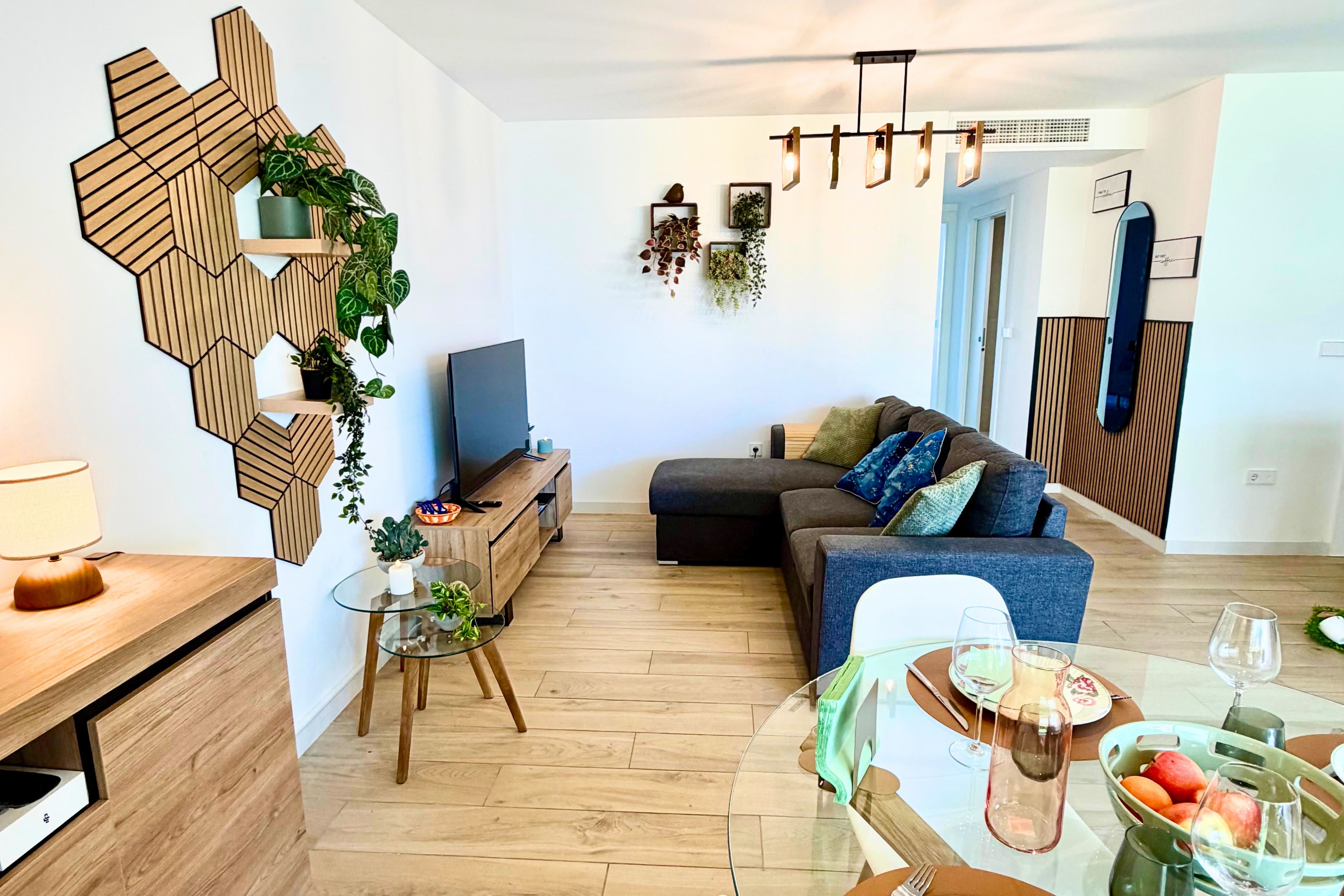Una sala de estar moderna con una decoración geométrica de madera, un sofá con cojines, una mesa de centro de vidrio con fruta y un televisor sobre un mueble de madera.