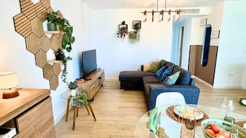 Una sala de estar moderna con una decoración geométrica de madera, un sofá con cojines, una mesa de centro de vidrio con fruta y un televisor sobre un mueble de madera.