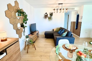 A modern living room with a wooden geometric wall decoration, a sofa with cushions, a glass coffee table with fruit, and a TV on a wooden stand. - Casa Rosalia 2 by Rental Hero (Alcazares, Los)