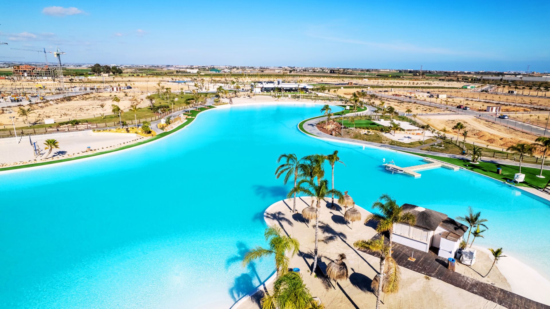 Una amplia piscina azul cristalina con palmeras y una zona de playa arenosa, rodeada de un paisaje cuidado con edificios y carreteras.