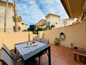 An outdoor patio with a table set for two, surrounded by chairs, and a small garden area with a palm tree and a potted plant. - Casa Brisa Mediterranea by Rental Hero (Alcazares, Los)