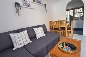 A modern living room with a grey sofa, two patterned cushions, a wooden coffee table with remote controls, and a dining area with wooden chairs and a table. - Casa Margarita by Rental Hero (Alcazares, Los)
