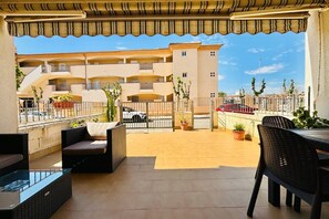 A covered outdoor area with a seating arrangement, a glass-top table, and a view of a building with multiple balconies. - Casa Abuelo Fico by Rental Hero (Alcazares, Los)