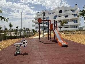 A playground with a slide and climbing structures, surrounded by a paved area with a soccer ball and a skateboard. - Casa Emerald 2 by Rental Hero (Alcazares, Los)