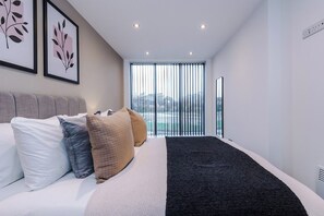 Ein modernes Schlafzimmer mit Bett, Kissen, einem schwarzen Teppich und einem großen Fenster mit Jalousien.