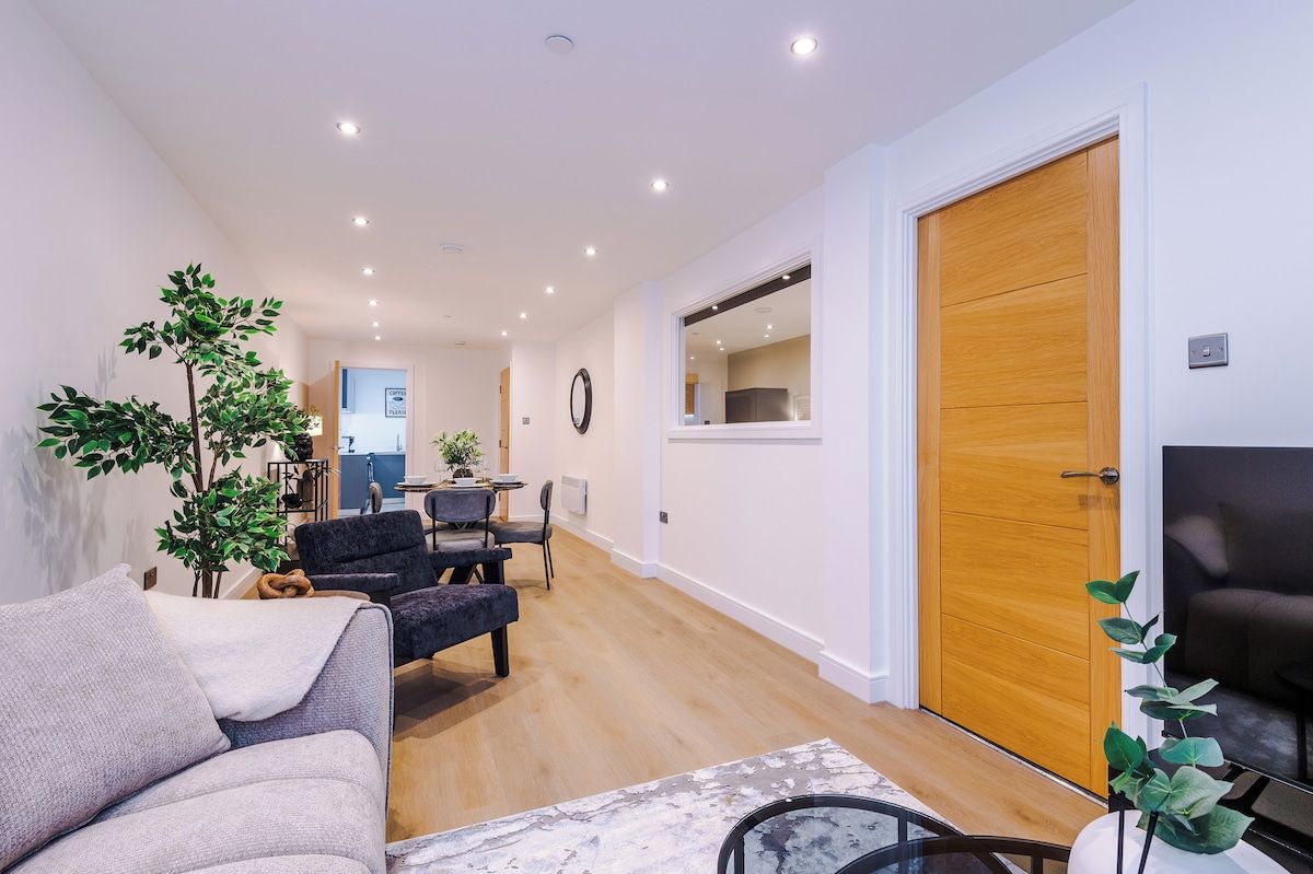 Una sala moderna con sofá, sillones, una mesa de centro y televisor. Hay una puerta de madera y una ventana con vistas.