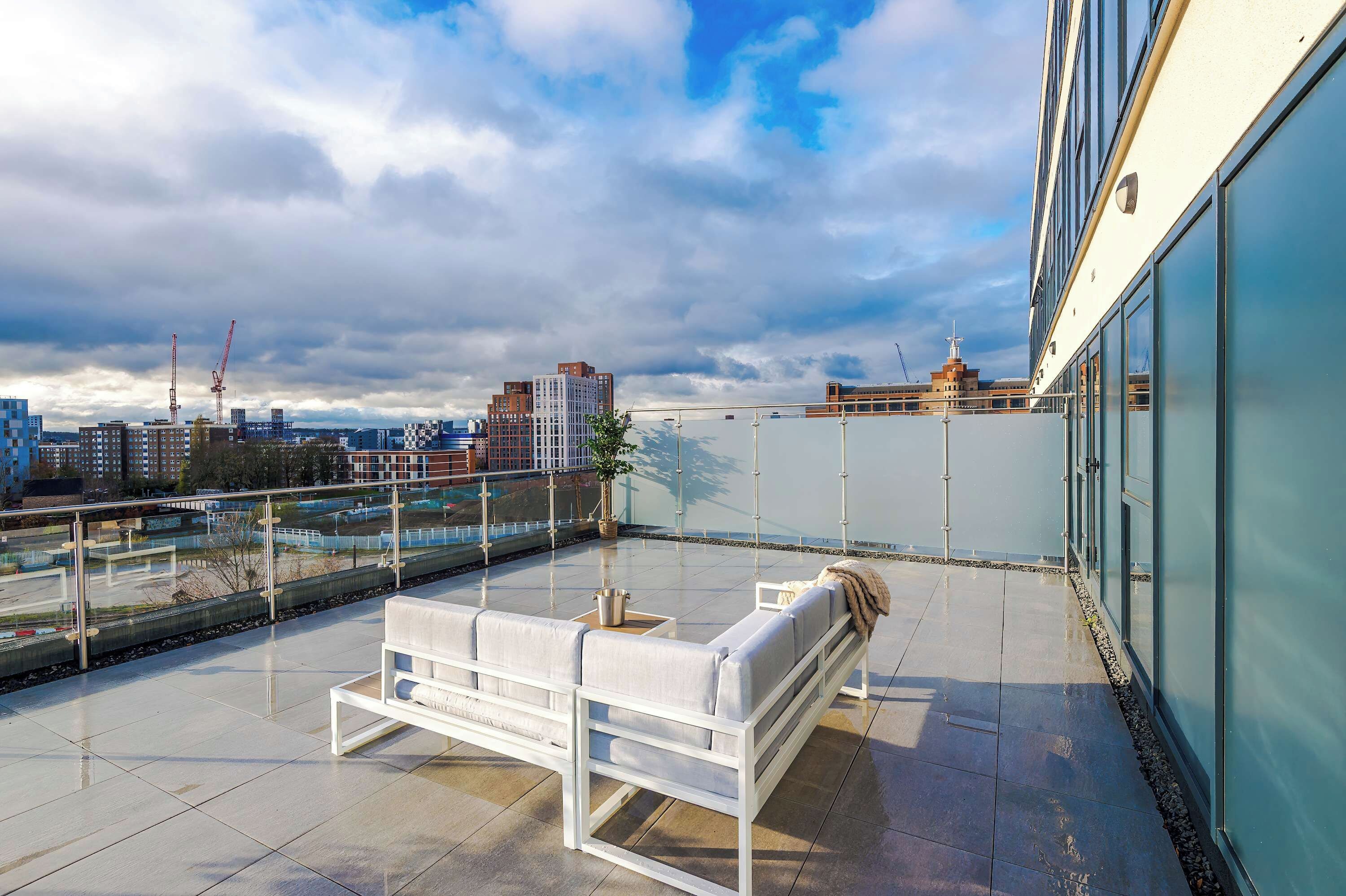 A rooftop terrace with a white sofa and a small table, overlooking a cityscape with buildings and a construction crane.