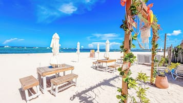 Een strandsetting met houten tafels en stoelen, parasols en een houten omheining versierd met planten.