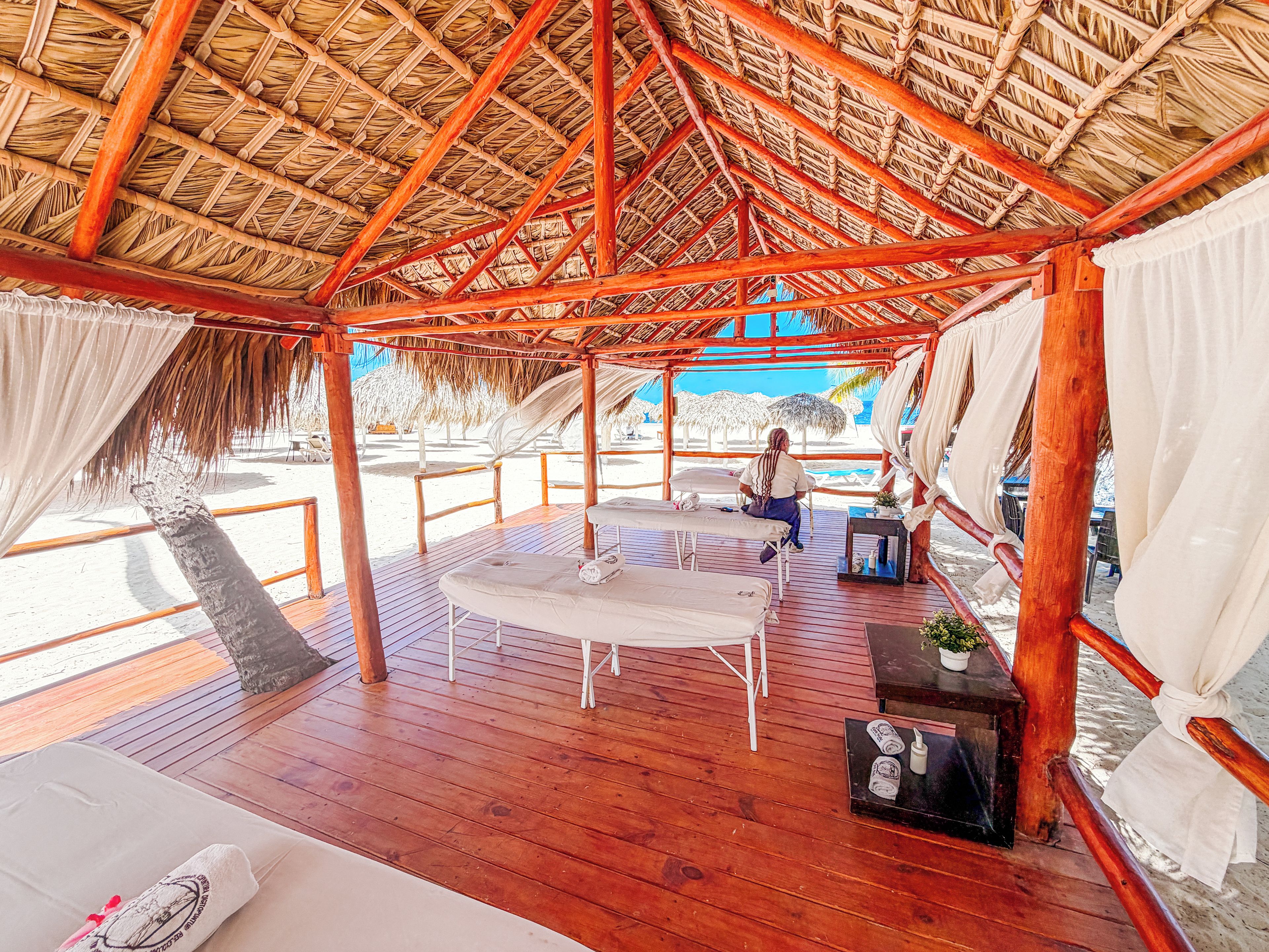 Un espacio al aire libre cubierto con una terraza de madera, mobiliario blanco y una estructura de techo de paja.
