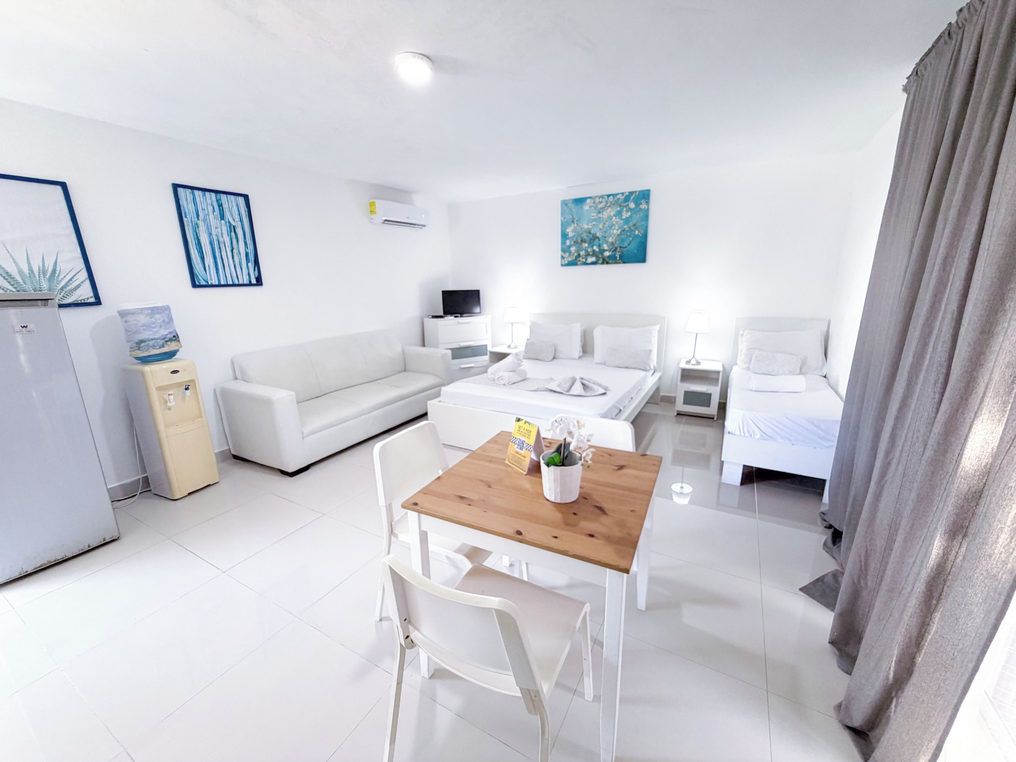 Una habitación de hotel moderna con cama, mesitas de noche, sofá, mesa de comedor con sillas, dispensador de agua y obras de arte en las paredes.