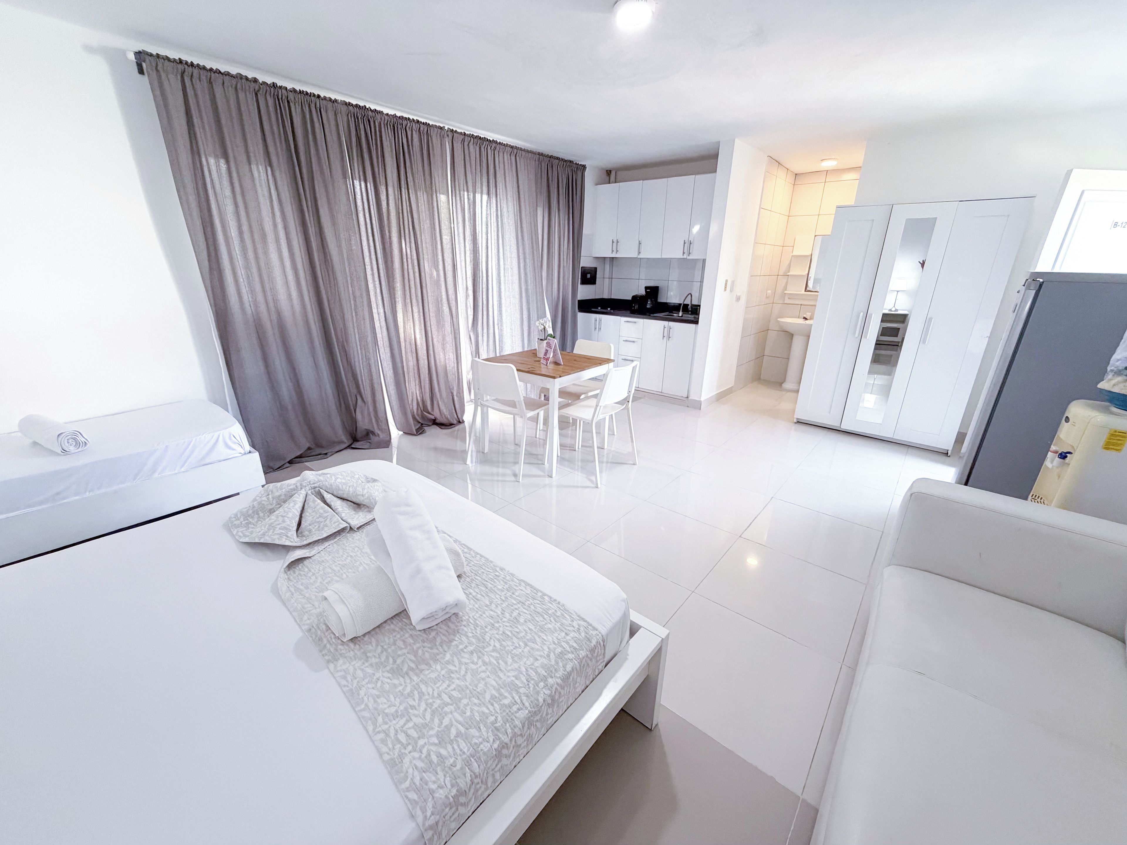 Una habitación de hotel moderna con una cama, una mesita y una kitchenette al fondo.