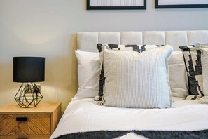 A neatly made bed with a white duvet and multiple pillows, a bedside table with a lamp, and a wooden nightstand with drawers. - City Centre Flat with a Terrace (West Yorkshire)