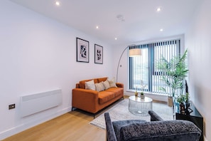 A modern living room with a sofa, a coffee table, and a floor lamp. There are two framed artworks on the wall and a large window with blinds. - City Centre Flat with a Terrace (West Yorkshire)