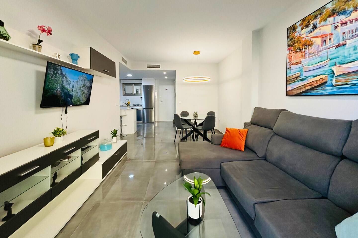A modern living room with a grey sofa, a flat-screen TV, a dining area with a table and chairs, and a kitchen in the background.