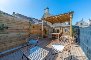 Eine Holzterrasse mit Gartenmöbeln, einer Pergola und einem Steinhaus im Hintergrund.
