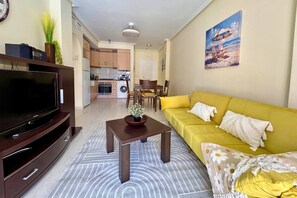 A living room with a flat-screen TV, a yellow sofa, a coffee table, and a kitchen in the background. - Casa Pool Paradise by Rental Hero (Alcazares, Los)
