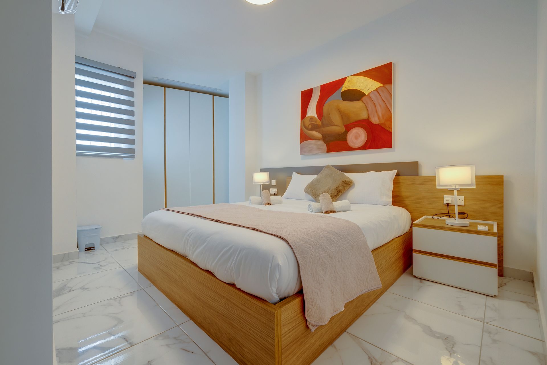 Un dormitorio moderno con una cama de madera, ropa de cama blanca, un plaid color beige, dos mesitas de noche con lámparas y un cuadro abstracto grande en la pared.
