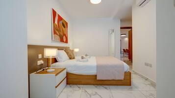 Una habitación de hotel moderna con una cama de madera, ropa de cama blanca, una mesita de noche con lámpara y un cuadro en la pared.