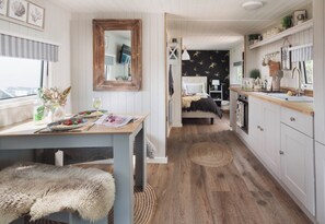 Ein gemütliches Interieur mit einem Holzesstisch, einer Bank, einem Bett mit weißem Kopfteil und einer Küchenzeile mit weißen Schränken und einer Holz Arbeitsplatte.