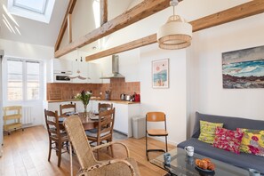 A modern kitchen with wooden beams, a dining area with a glass table, and a living room with a sofa and colorful cushions. - Le Macareux - Duplex pour 4 (Saint-Malo)