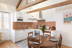 A modern kitchen with white cabinets, a wooden dining table set for four, and a painting on the wall. - Le Macareux - Duplex pour 4 (Saint-Malo)