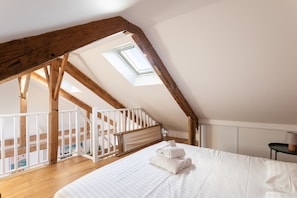 A bedroom with a sloped ceiling, wooden beams, a skylight, a bed with white bedding, and a view of a balcony. - Le Macareux - Duplex pour 4 (Saint-Malo)