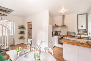A modern kitchen with white cabinetry, a wooden countertop, and a built-in oven. There is a dining area with a white table and chairs, and a staircase leading to an upper level. - Le Cocon du Bel Air - 500m de la Plage (Saint-Malo)