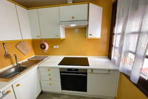 A compact kitchen with white cabinets, a sink, a stove, and a window with curtains. - Casa Rio Nalon by Rental Hero (Alcazares, Los)