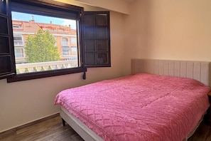 A bedroom with a bed, a window with a view of buildings, and a wooden floor. - Casa Rio Nalon by Rental Hero (Alcazares, Los)