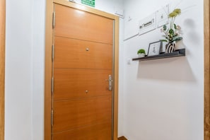Uma porta de madeira com maçaneta e uma pequena fechadura, uma prateleira fixada na parede com objetos decorativos e uma placa de saída de emergência verde acima da porta.