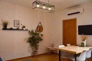 A modern dining area with a wooden table, white chairs, a small side table, a potted plant, a wall-mounted mirror, and a ceiling light fixture. - Apartment near Plaza de Espana, Madrid (Madrid)