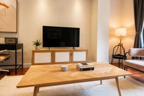 Ein modernes Wohnzimmer mit einem großen Flachbildfernseher auf einer Holzkommode, einem Couchtisch mit Büchern und einer Stapel Untersetzer, einer Stehlampe und einem Sofa.