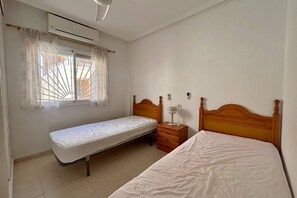A bedroom with two beds, a window with curtains, an air conditioning unit, and a bedside table with a lamp. - Casa San Juda by Rental Hero (Alcazares, Los)