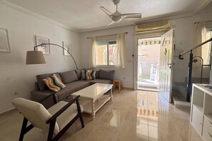 A living room with a sofa, coffee table, and armchair, a ceiling fan, and a view through a door to the outside. - Casa San Juda by Rental Hero (Alcazares, Los)