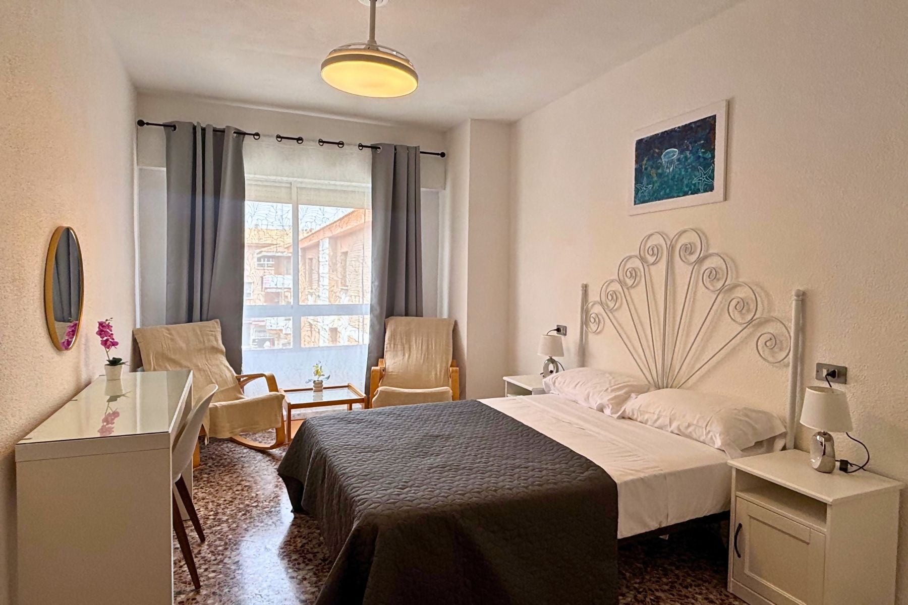 A hotel room with a bed, two chairs, a desk, a lamp, and a painting on the wall.