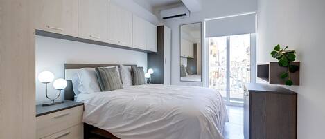 Un dormitorio moderno con una cama grande, lámparas de noche, un aparador, un espejo y vistas de la ciudad a través de un ventanal.