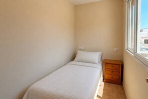 A small, single-bed room with a wooden nightstand and a window providing a view of buildings outside. - Casa de la Luz by Rental Hero (Alcazares, Los)