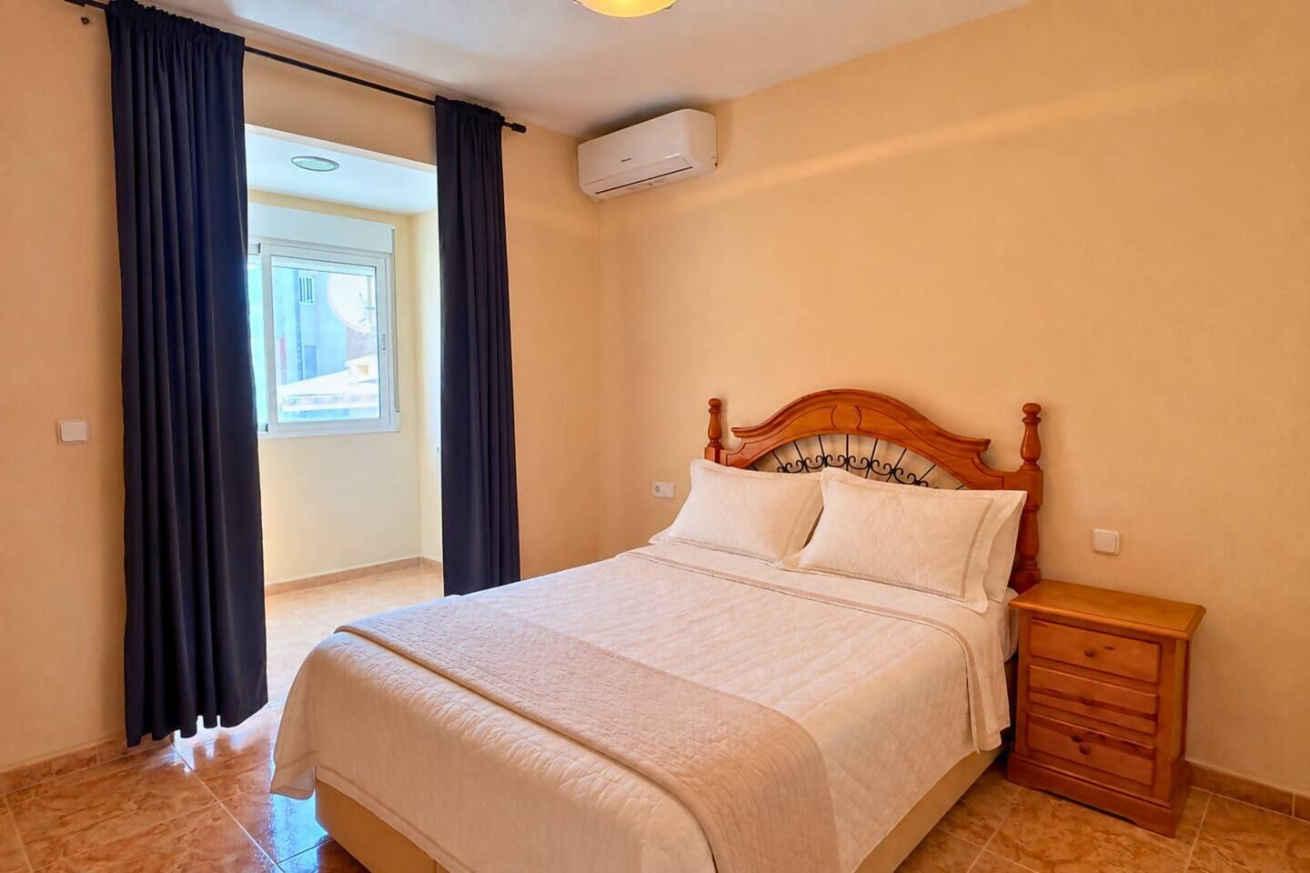 Une chambre à coucher comprenant un lit, une table de chevet, une fenêtre avec des rideaux et un appareil de climatisation.