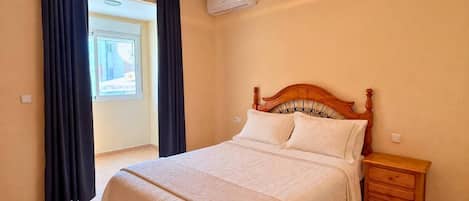 A bedroom with a bed, a nightstand, a window with curtains, and an air conditioning unit.