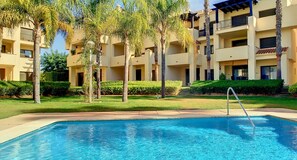 A swimming pool with a metal ladder, surrounded by well-maintained grass and palm trees, with a multi-story building in the background. - Casa Roda Golf by Rental Hero (Roda)