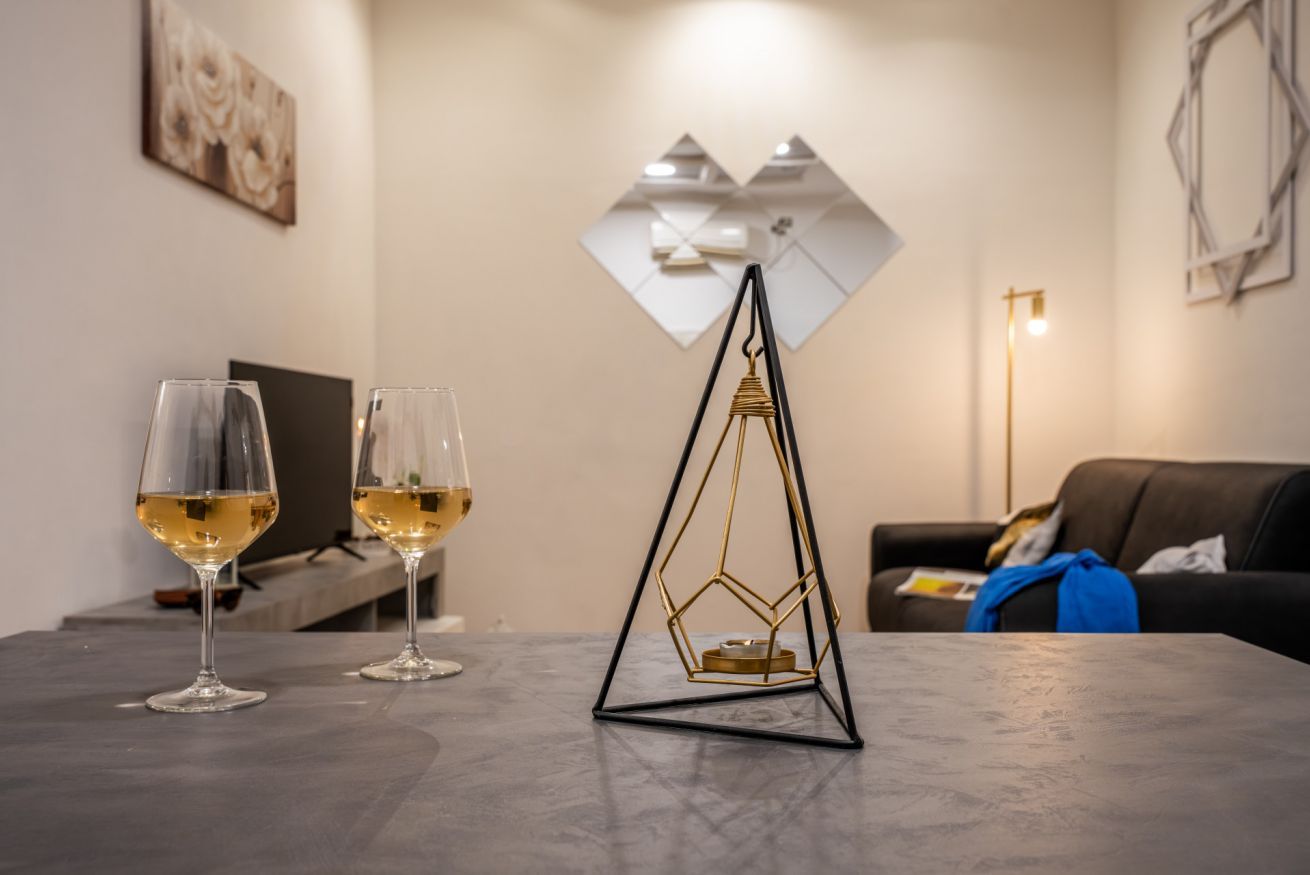 Un salon moderne avec une table en verre, deux verres à vin, un objet décoratif aux motifs géométriques et un canapé recouvert d’une couverture.