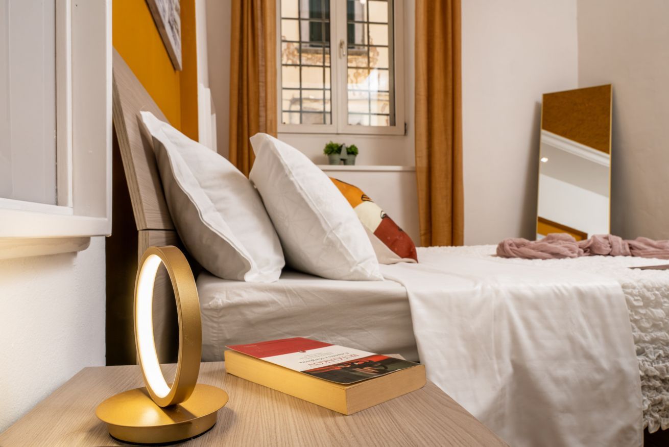 Un lit bien fait, avec des oreillers, une table de chevet sur laquelle se trouvent un livre et une lampe, et une fenêtre avec des rideaux.
