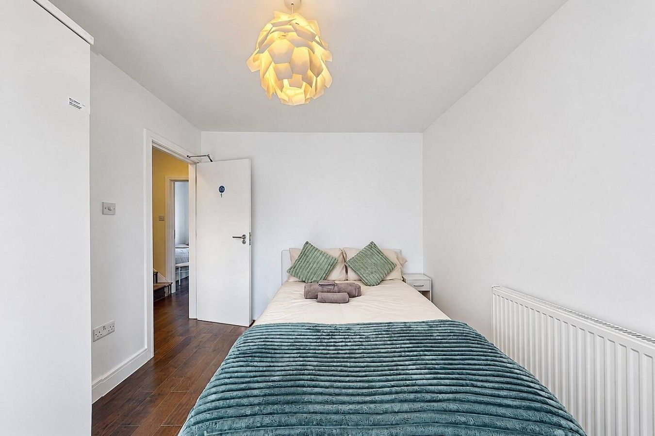 A bedroom with a bed, bedside tables, a radiator, and a door leading to another room.