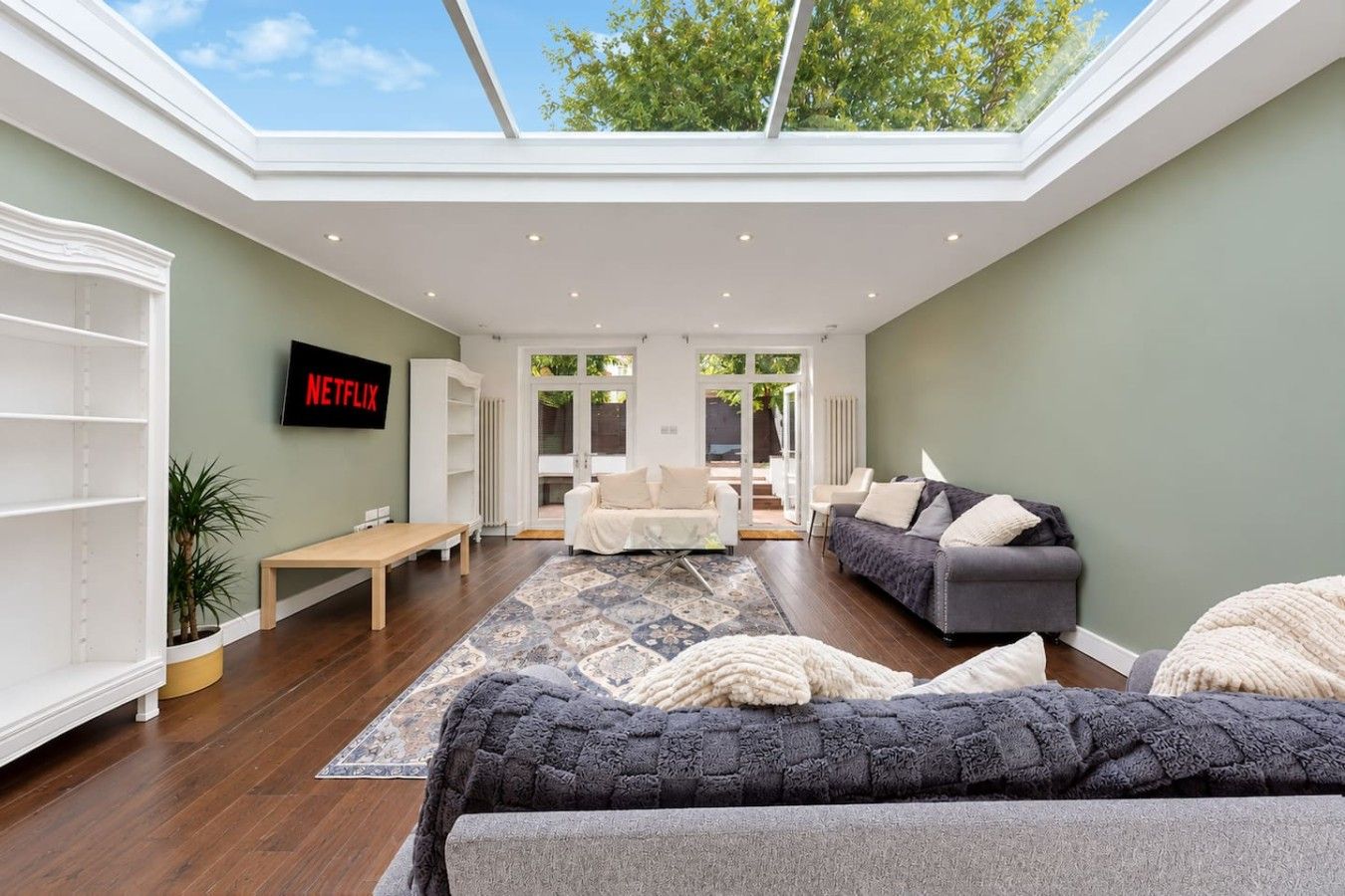 A modern living room with a high ceiling, a flat-screen TV, a sofa, a coffee table, and a view through a glass door to the outside.