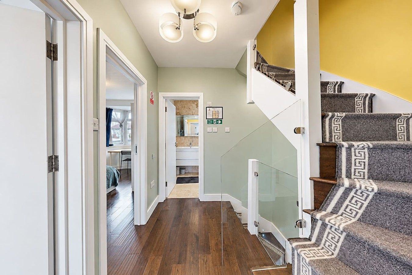 A hallway with a wooden floor, a glass railing staircase, and a door leading to another room.