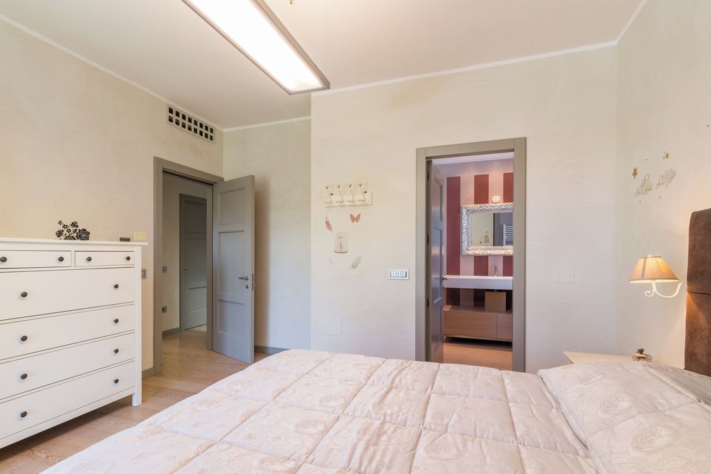 Un dormitorio con cama, cómoda, puerta que da a un baño y una lámpara fijada a la pared.