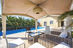 A covered outdoor seating area with a pool, wicker furniture, and a stone building in the background.
